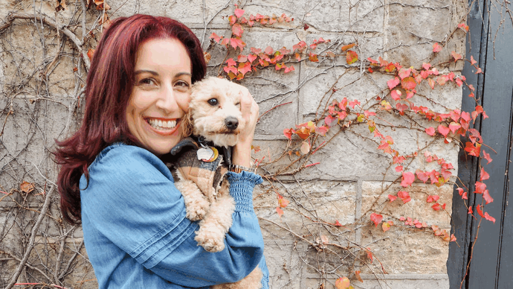 Woman smiling and holding a small dog beside an ivy-covered stone wall.